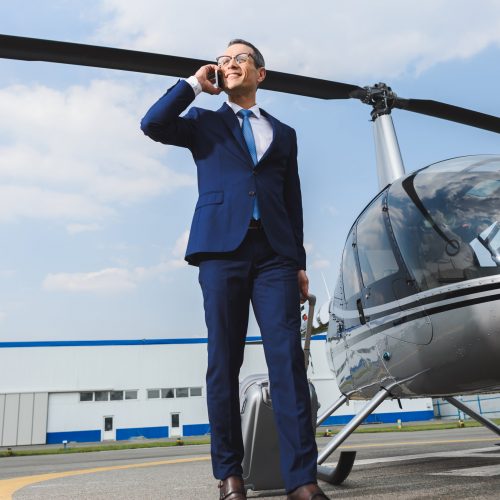 businessman in formal wear with suitcase talking on smartphone near helicopter